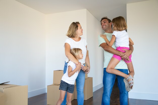 Heureux parents et enfants appréciant de déménager dans un nouvel appartement, debout près de la pile de boîtes et étreindre