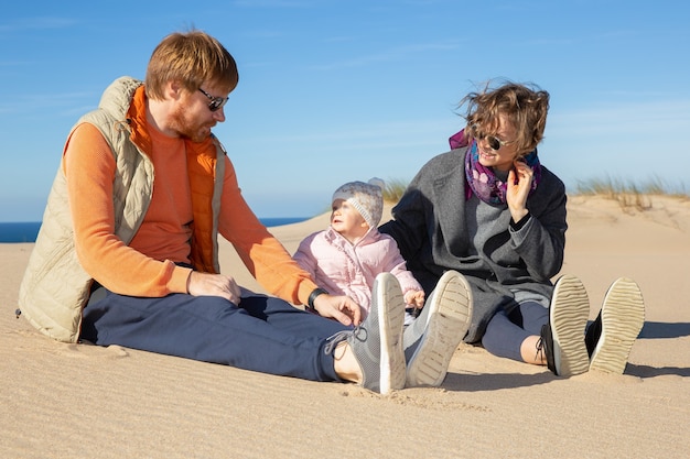 Heureux parents et jolie petite fille portant des vêtements chauds, profitant du temps libre en mer, assis sur le sable ensemble