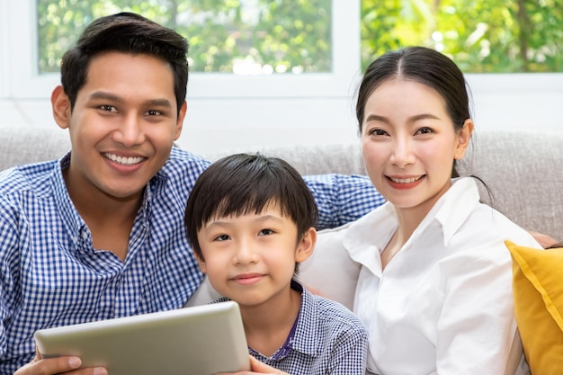 Heureux père de famille asiatique mère et fils assis sur un canapé en utilisant une tablette de jeu ensemble dans le salon en regardant la caméra