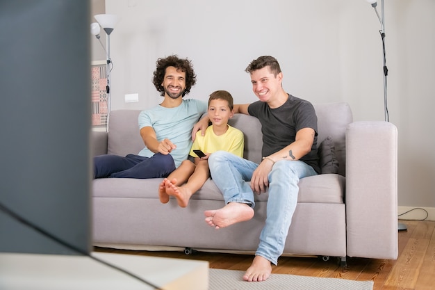 Heureux pères et fils gay regardant une émission de télévision drôle à la maison, assis sur un canapé dans le salon, souriant et riant. Concept de divertissement familial et à domicile