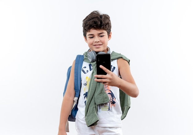 Heureux petit écolier portant sac à dos et écouteurs tenant et regardant le téléphone isolé sur fond blanc