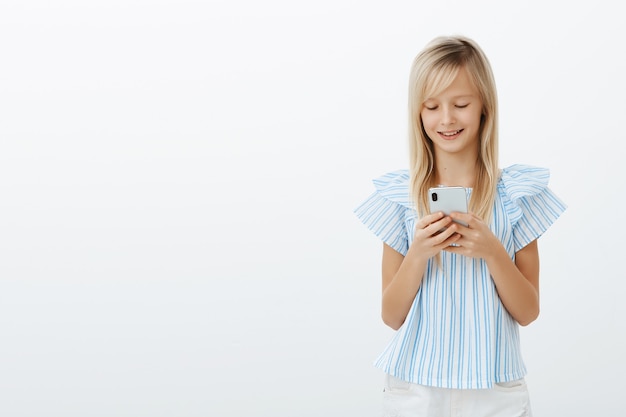 Heureux petit enfant de sexe féminin aux cheveux blonds en chemisier bleu, tenant un smartphone et souriant à l'écran tout en envoyant des messages ou en jouant à un jeu amusant