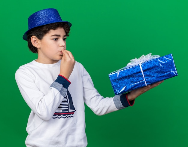 Heureux petit garçon portant un chapeau de fête bleu tenant et regardant une boîte-cadeau montrant un geste délicieux