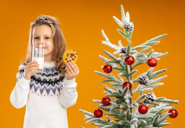 Heureux petite fille debout à proximité de l'arbre de Noël portant diadème avec guirlande sur le cou tenant un verre de lait avec des biscuits isolé sur fond orange
