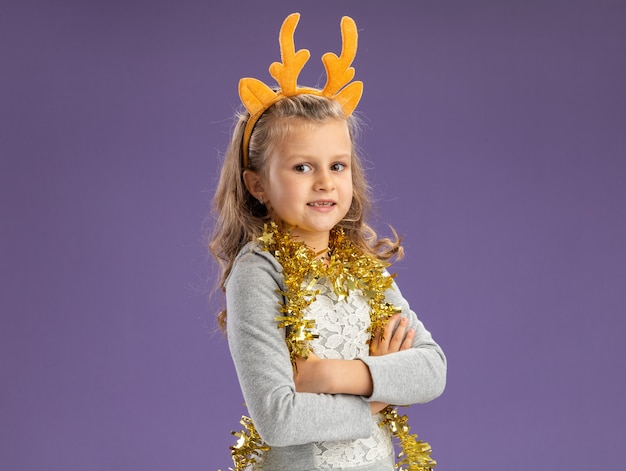 Heureux petite fille portant un cerceau de cheveux de Noël avec guirlande sur le cou croisant les mains isolé sur fond bleu