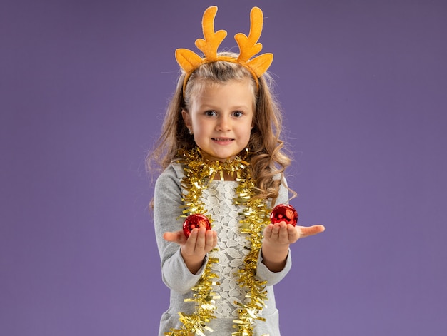 Heureux petite fille portant un cerceau de cheveux de Noël avec guirlande sur le cou tenant des boules de Noël isolé sur fond bleu