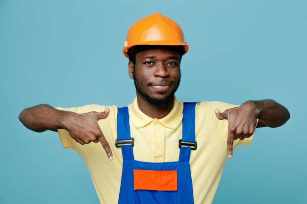 heureux pointe vers le bas jeune constructeur afro-américain en uniforme isolé sur fond bleu