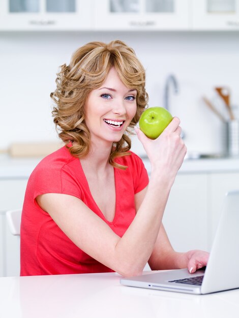 Heureux portrait de belle jeune femme assise dans la cuisine et manger une pomme verte