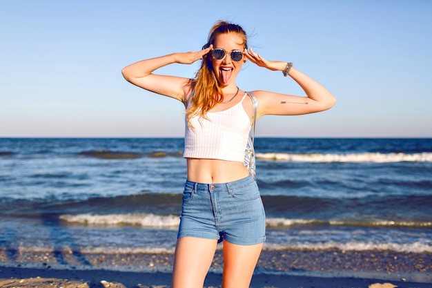 Heureux portrait positif de jeune femme blonde s'amusant à des vacances tropicales, marchant près de l'océan, voyage avec sac à dos, tenue d'été hipster élégante et lunettes de soleil. corps sexy bronzé, poils longs.