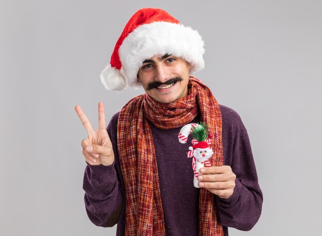 Heureux et positif jeune homme moustachu portant bonnet de noel de Noël avec écharpe chaude autour de son cou tenant la canne de Noël souriant montrant v-sign