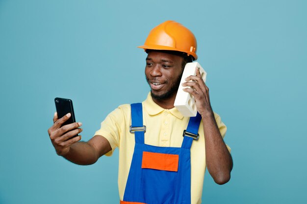 Heureux de prendre un selfie mettant de la brique sur l'oreille jeune constructeur afro-américain en uniforme isolé sur fond bleu