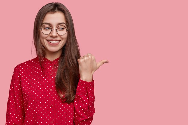 Heureux le promoteur a un sourire radieux, montre du pouce un espace vide, vêtu de vêtements élégants rouges