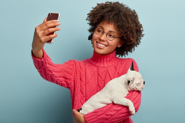 Heureux propriétaire de chien incline la tête, a une coiffure afro, vêtu d'un pull rose, tient un petit chiot endormi