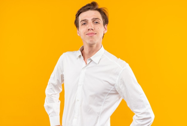 Heureux à la recherche d'appareil photo jeune beau mec vêtu d'une chemise blanche mettant les mains sur la hanche isolé sur le mur orange