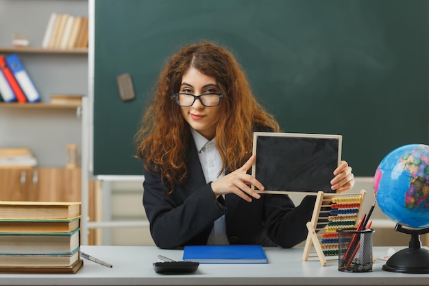 heureux regardant la caméra jeune enseignante portant des lunettes tenant un mini tableau assis au bureau avec des outils scolaires en classe