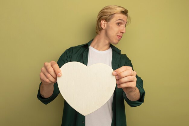 Heureux regardant côté jeune mec blond portant un t-shirt vert tenant une boîte en forme de coeur