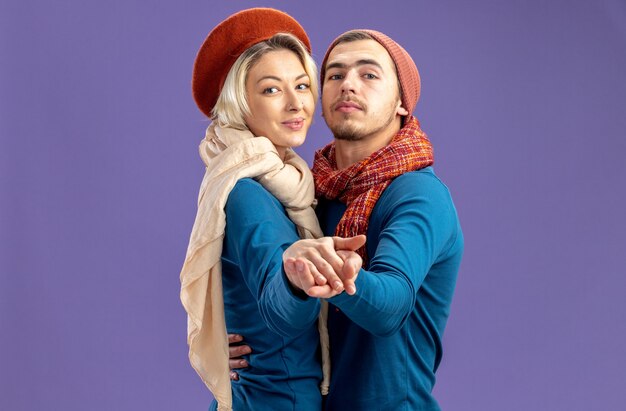 Heureux de regarder la caméra jeune couple portant un chapeau avec une écharpe le jour de la danse de la Saint-Valentin isolé sur fond bleu