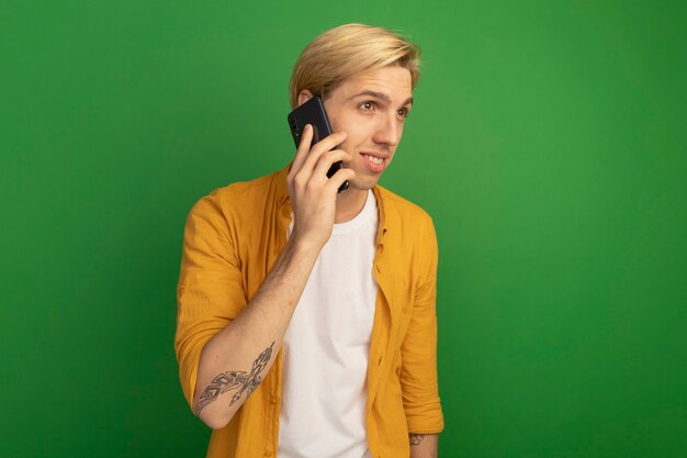 Heureux de regarder à côté jeune mec blond portant un t-shirt jaune parle au téléphone isolé sur vert avec copie espace