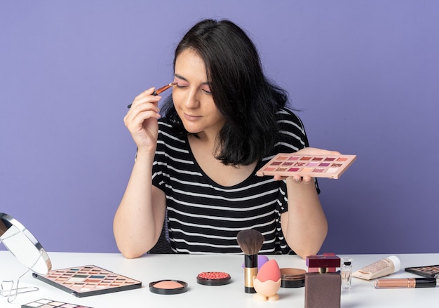 Heureux de regarder le miroir jeune belle fille assise à table avec des outils de maquillage appliquant un fard à paupières avec un pinceau de maquillage isolé sur un mur bleu