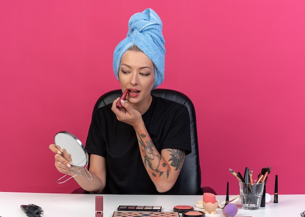 Heureux de regarder miroir jeune belle fille assise à table avec des outils de maquillage cheveux enveloppés dans une serviette appliquant du rouge à lèvres isolé sur mur rose