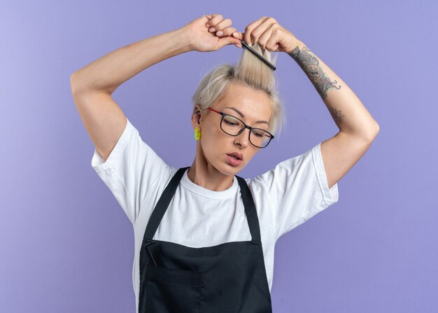 Heureux de regarder vers le bas la belle jeune femme barbier en uniforme portant des lunettes peignant les cheveux isolés sur le mur bleu