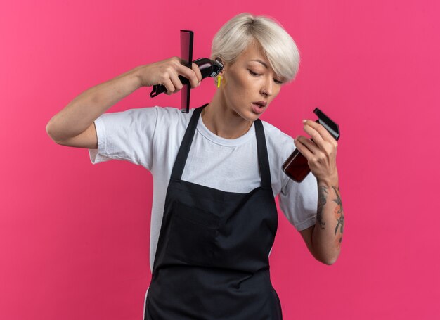 Heureux de regarder vers le bas une belle jeune femme coiffeuse en uniforme tenant des outils de coiffeur isolés sur un mur rose