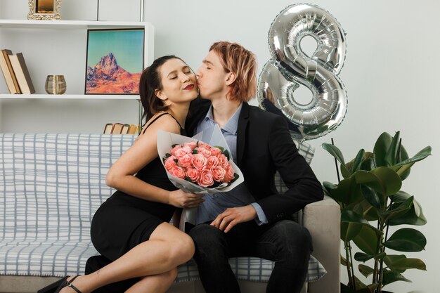 Heureux de s'embrasser jeune couple lors de la journée des femmes heureuses girl holding bouquet sitting on sofa in living room