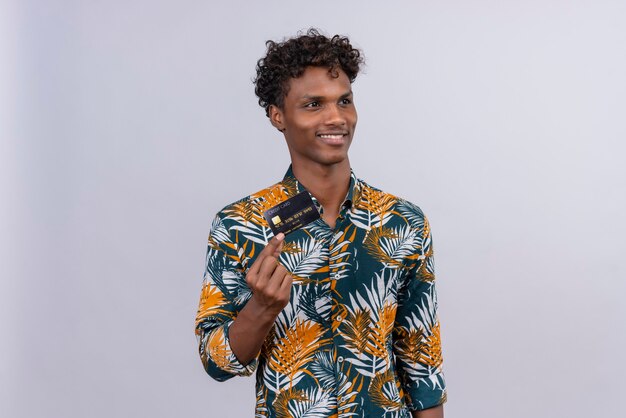 Heureux et souriant bel homme à la peau sombre avec des cheveux bouclés en chemise imprimée de feuilles montrant la carte de crédit