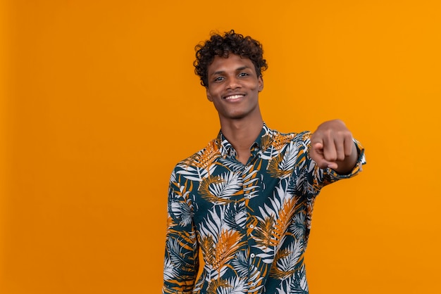 Heureux et souriant jeune bel homme à la peau foncée aux cheveux bouclés en chemise imprimée de feuilles tout en pointant avec le doigt