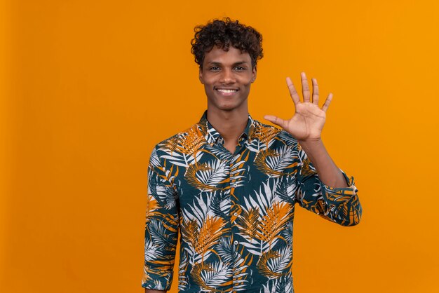 Heureux et souriant jeune bel homme à la peau foncée avec des cheveux bouclés en chemise imprimée de feuilles tout en montrant avec les doigts numéro cinq