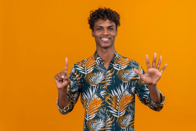 Heureux et souriant jeune bel homme à la peau foncée avec des cheveux bouclés en chemise imprimée de feuilles tout en montrant avec les doigts numéro six