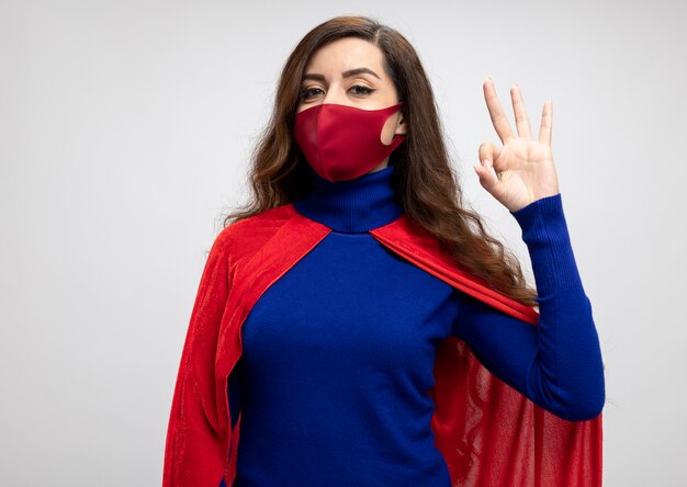 Heureux superwoman avec cape rouge portant des gestes de masque de protection rouge ok signe de la main isolé sur mur blanc