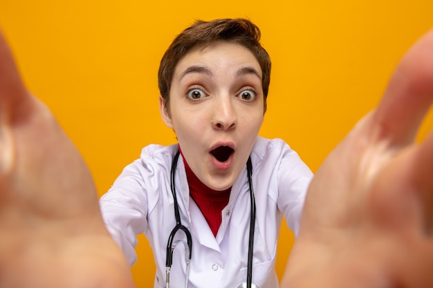 Heureux et surpris jeune fille médecin en blouse blanche avec stéthoscope autour du cou prenant selfie sur orange