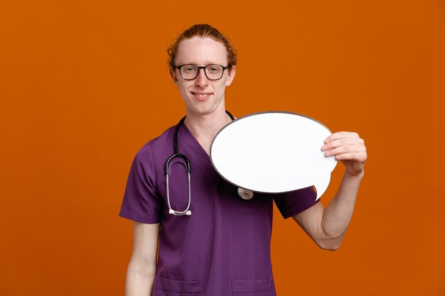 Heureux tenant bulle jeune homme médecin portant l'uniforme avec stéthoscope isolé sur fond orange