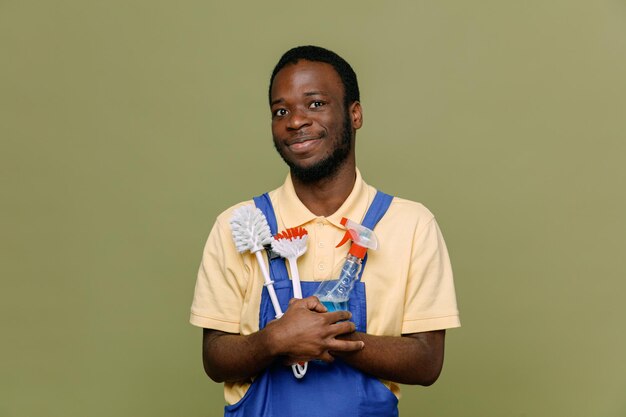 Heureux tenant des outils de nettoyage jeune homme nettoyant afro-américain en uniforme avec des gants isolés sur fond vert