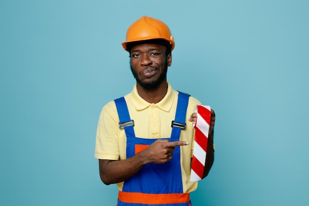 Heureux tenant et points ruban adhésif jeune constructeur afro-américain en uniforme isolé sur fond bleu