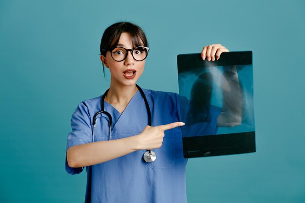 Heureux tenant et points à xray jeune femme médecin portant un stéthoscope fith uniforme isolé sur fond bleu