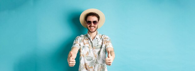 Heureux touriste en lunettes de soleil et chapeau de paille montrant les pouces vers le haut et souriant profitant des vacances d'été stan