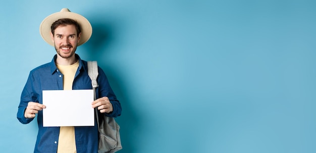 Photo gratuite heureux touriste avec sac à dos montrant un morceau de papier vide souriant à la caméra debout sur le backgrou bleu