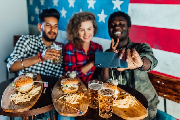 Photo gratuite heureux trois amis dans un restaurant de restauration rapide prenant un selfie pendant qu'ils mangent des hamburgers et boivent de la bière