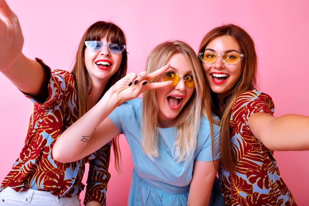 Photo gratuite heureux trois filles élégantes souriantes s'amusant, montrant le geste de paix et souriant, lunettes de soleil hipster à la mode et vêtements assortis à la mode, objectifs d'amitié, mur rose