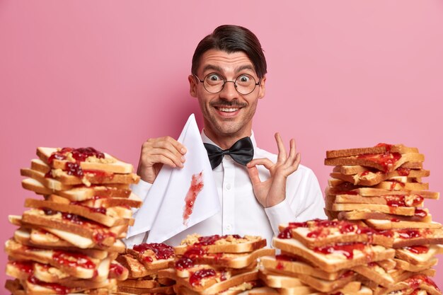 Heureux visiteur de café homme élégant ajuste le nœud papillon, ayant faim et prêt à manger de délicieux toasts avec de la confiture, tient une serviette, a des manières polies, isolé sur un mur rose. Gens, concept de manger