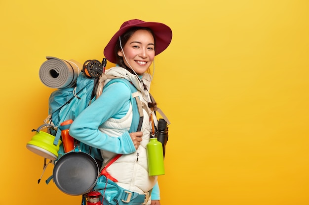 Heureux voyageur asiatique brune porte un grand sac à dos touristique, utilise des jumelles pour le voyage, se tient contre le mur jaune, porte un chapeau élégant, un pull avec gilet