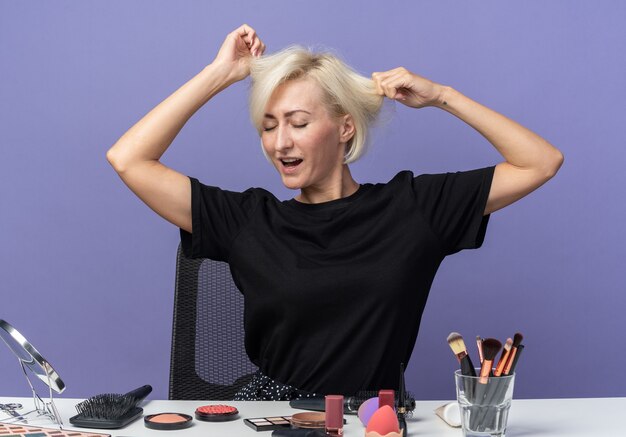 Heureux avec les yeux fermés, une belle jeune fille est assise à table avec des outils de maquillage attrapé les cheveux isolés sur le mur bleu