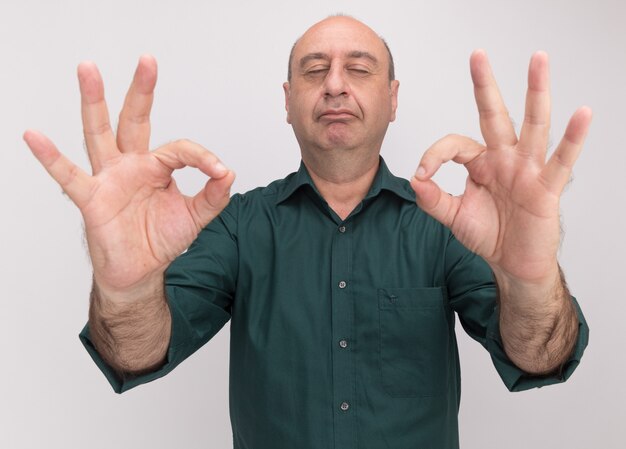 Heureux avec les yeux fermés homme d'âge moyen portant un t-shirt vert faisant de la méditation isolé sur mur blanc