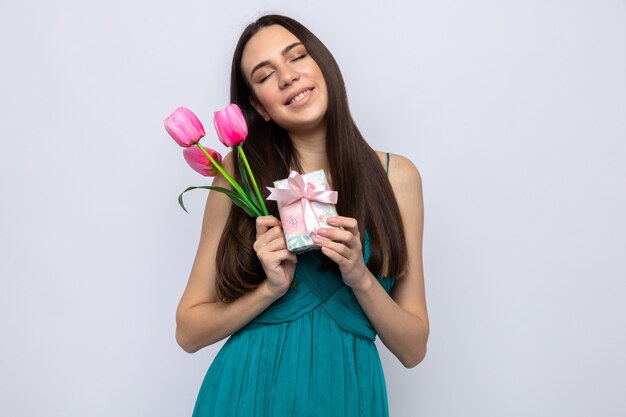 Heureux avec les yeux fermés inclinant la tête belle jeune fille le jour de la femme heureuse tenant un cadeau avec des fleurs isolées sur un mur blanc