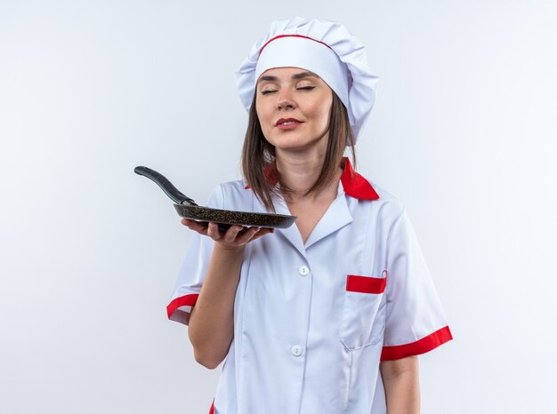 Heureux avec les yeux fermés jeune cuisinière portant un uniforme de chef tenant une poêle à frire isolée sur un mur blanc