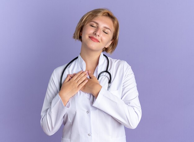 Heureux avec les yeux fermés jeune femme médecin portant une robe médicale avec stéthoscope mettant les mains sur elle-même isolée sur le mur bleu