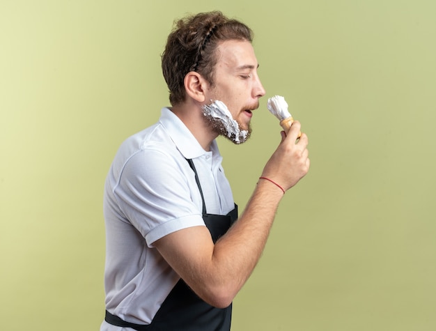 Heureux avec les yeux fermés jeune homme barbier portant une crème à raser appliquée uniforme avec blaireau isolé sur mur vert olive