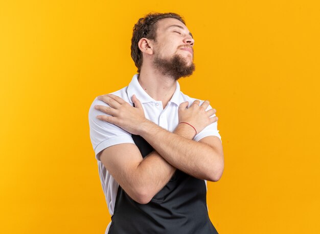 Heureux avec les yeux fermés jeune homme barbier en uniforme mettant les mains sur l'épaule isolé sur mur jaune